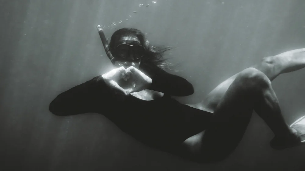 A black and white photograph of a guest snorkelling underwater, making a heart shape with her hands