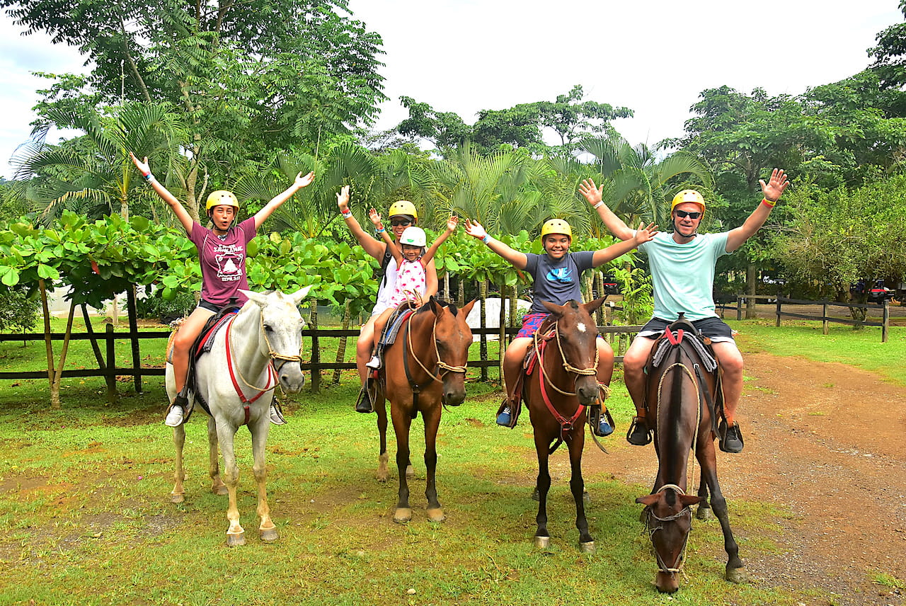 A family of four on horseback with their arms raised in the jungle