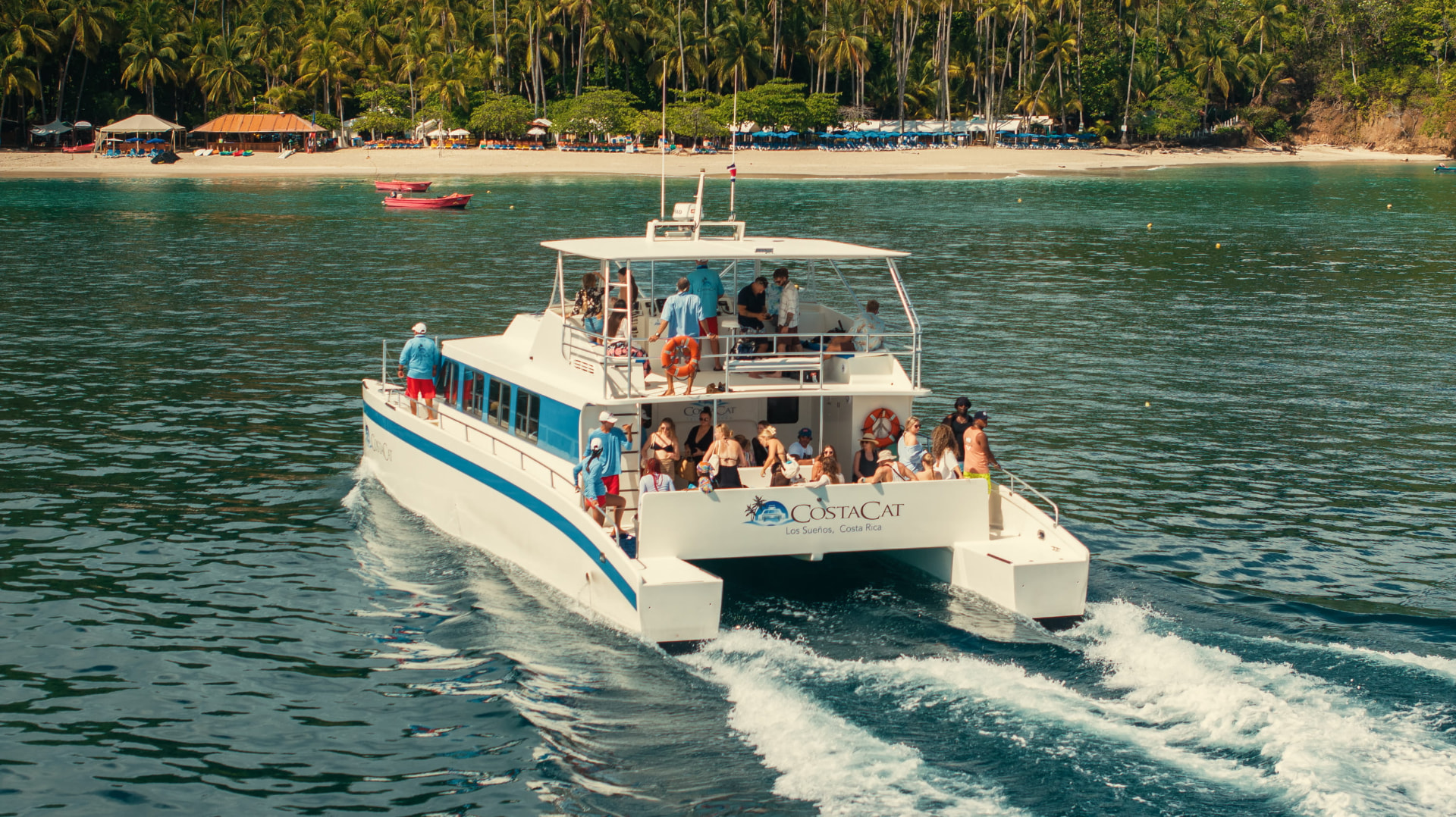 A catamaran full of guests cruising away from a palm-lined bay